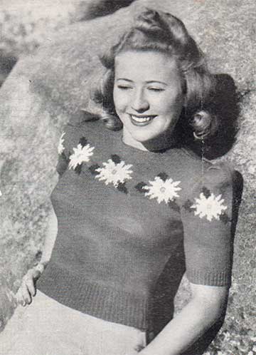 Woman in a close fitting knit top with crew neck, short puffed sleeves & a row of intarsia flowers across the chest & sleeves.