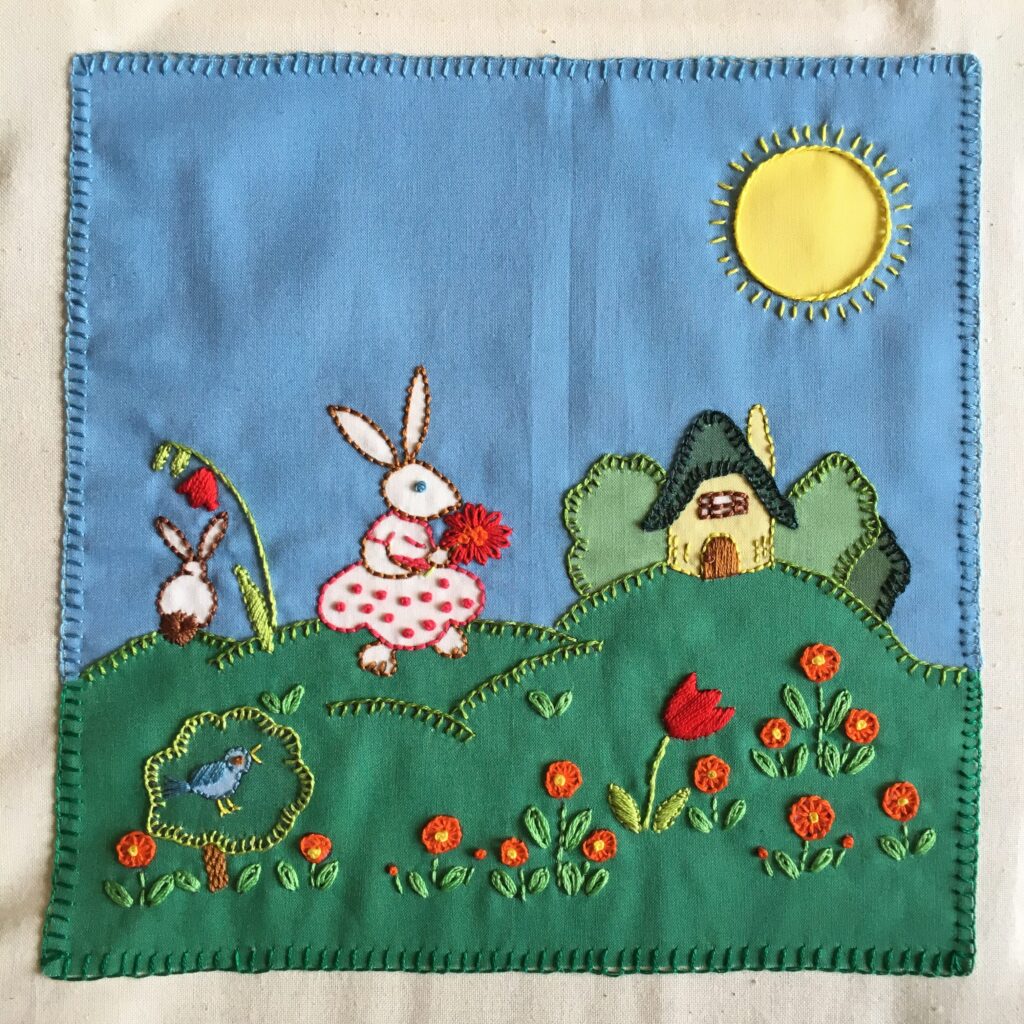 Embroidered square with two rabbits on a garden and a small house