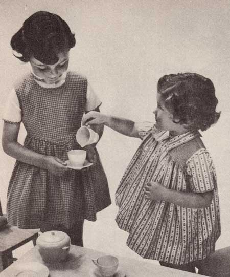 Two little girls wearing a pinafore and a gathered dress