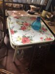 A small, white wicker side table with pink floral wallpaper on top.